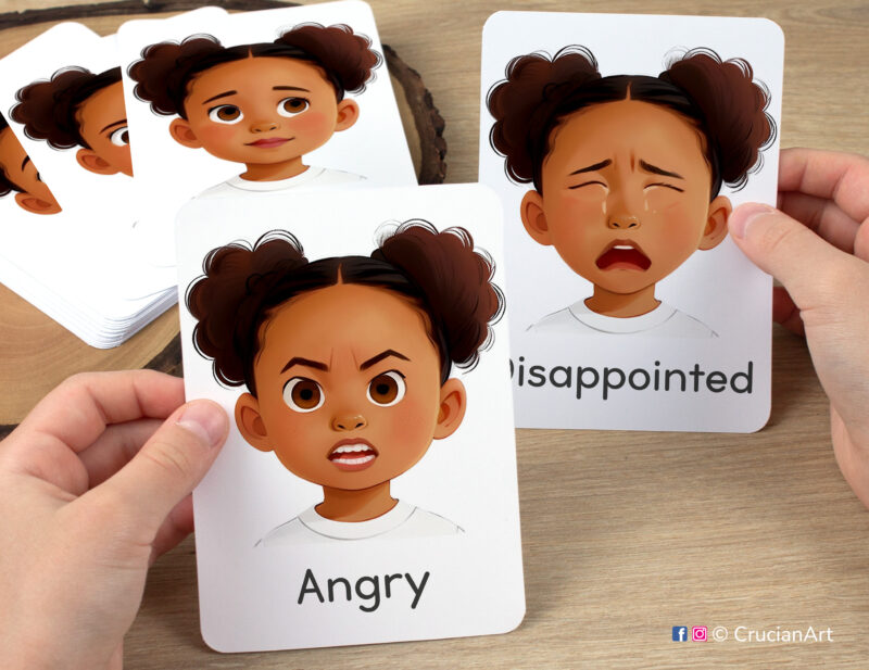 Preschooler hands holding flashcards with watercolor images of Angry and Disappointed girl faces. Emotions and Feelings flash cards for African American girls with brown skin and hair.