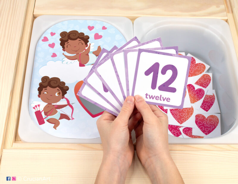 Pretend play setup for a Saint Valentine's Day counting game. Kids' hands holding task cards displaying numerals from 1 to 12.