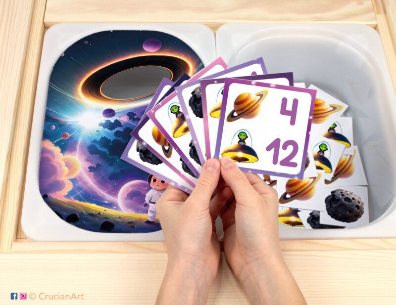 Outer Space unit pretend play setup for a matching and counting game. Kids' hands holding task cards displaying numerals and asteroids.