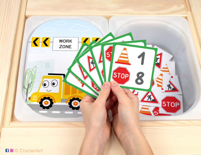 Construction Site theme pretend play setup for a matching and counting game. Kids' hands holding task cards displaying numerals and road signs.