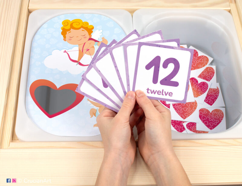 Pretend play setup for a Saint Valentine's Day counting game. Kids' hands holding task cards displaying numerals from 1 to 12.