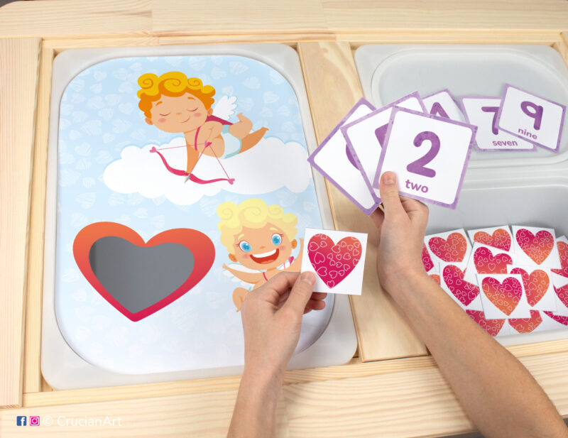 Toddler sensory play: Valentine's Day Cupid worksheet for a holiday educational activity inserted into IKEA Flisat table, with red hearts counters placed in the Trofast box.