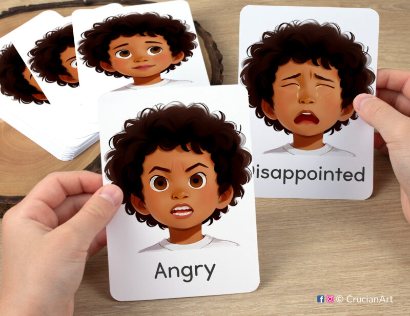 Preschooler hands holding flashcards with watercolor images of Angry and Disappointed boy faces. Emotions and Feelings flash cards for African American boys with brown skin and hair.