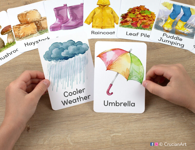 Preschooler's hands holding flashcards with watercolor images of Umbrella and Cooler Weather. Rainy season printable educational materials.