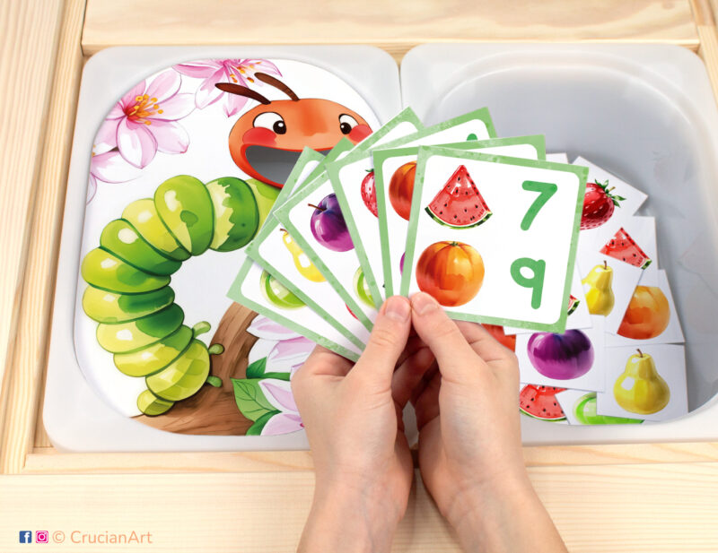 Very Hungry Caterpillar pretend play setup for a matching and counting game. Kids' hands holding task cards displaying numerals and fruits. Spring Insects unit printables for toddlers.
