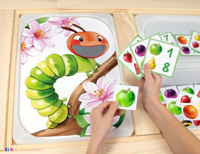 Toddler sensory play: Hungry Caterpillar worksheet for an educational activity inserted into IKEA Flisat table, with fruits counters with apples, plums, strawberries, watermelons, pears, oranges placed in the Trofast box.