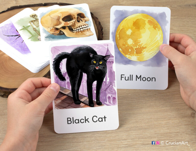 Preschooler hands holding flashcards with watercolor images of Black Cat and Full Moon. Set of fall season visual cards. Halloween Holiday printables.