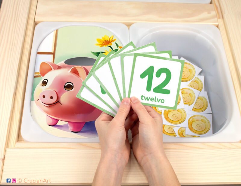 Piggy Bank pretend play setup for counting game. Sensory table insert and kids' hands holding task cards displaying numerals from 1 to 12.