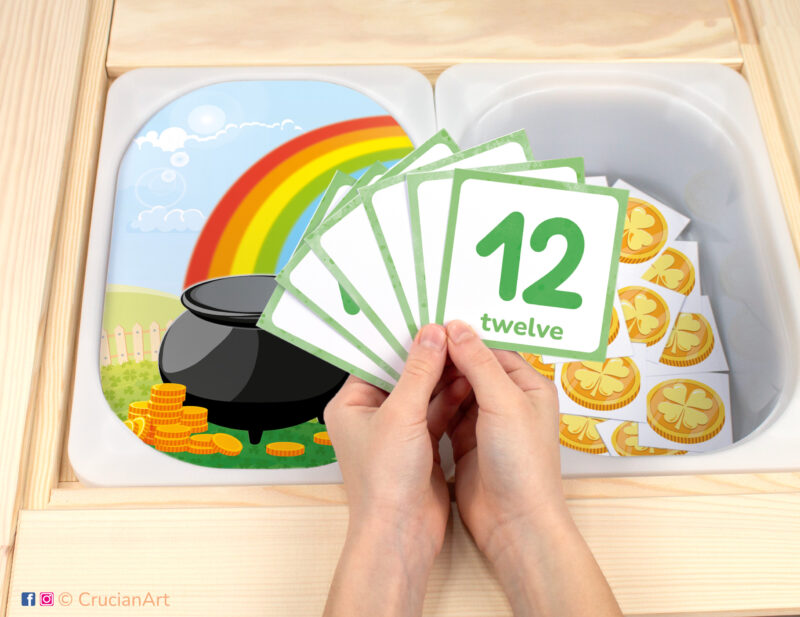 Leprechaun's Pot of Gold pretend play setup for a St. Patrick's Day theme counting game. Rainbow themed sensory table insert and kids' hands holding task cards displaying numerals from 1 to 12.