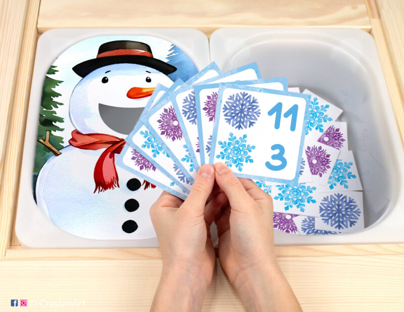 Winter unit pretend play setup for a matching and counting game. Kids' hands holding task cards displaying numerals and snowflakes.