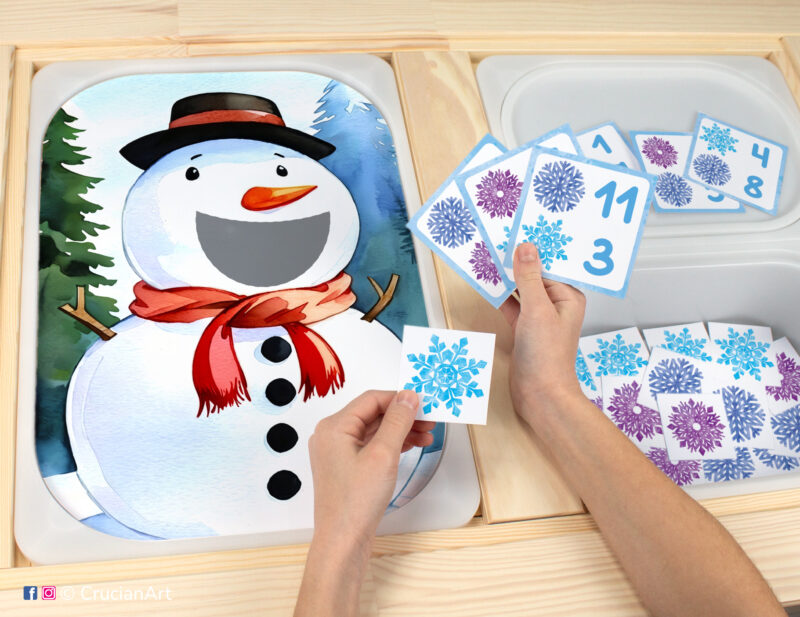 Toddler sensory play: Funny Snowman worksheet for an educational activity inserted into IKEA Flisat table, with snowflake counters placed in the Trofast box.