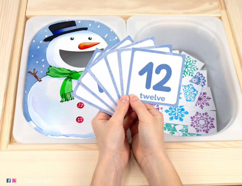 Pretend play setup for a winter counting game. Kids' hands holding task cards displaying numerals from 1 to 12.