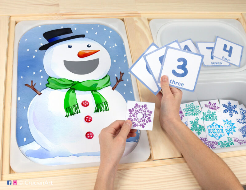 Toddler sensory play: Snowman worksheet for an educational activity inserted into IKEA Flisat table, with snowflake counters placed in the Trofast box.