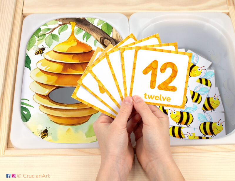 Beehive and wild bees pretend play setup for insect theme counting game. Honeybee themed sensory table insert and kids' hands holding task cards displaying numerals from 1 to 12.