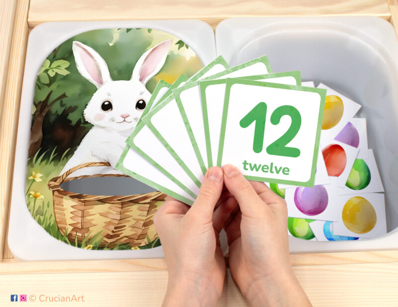 White bunny with basket pretend play setup for an Easter theme counting game. Easter bunny themed sensory table insert and kids' hands holding task cards displaying numerals from 1 to 12.