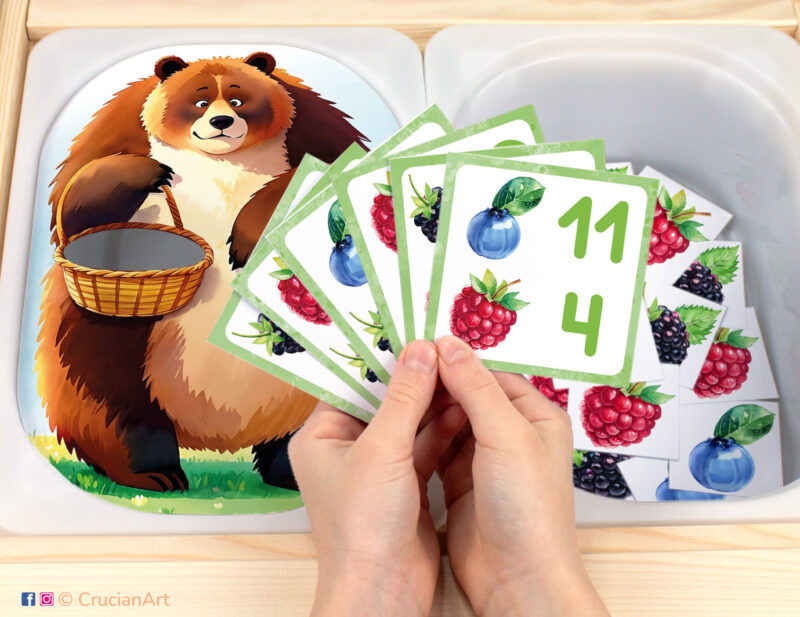 Feed the hungry brown bear berries pretend play setup for a matching and counting game. Kids' hands holding task cards displaying numerals and blueberries, raspberries, blackberries. Forest animals unit printables for toddlers.