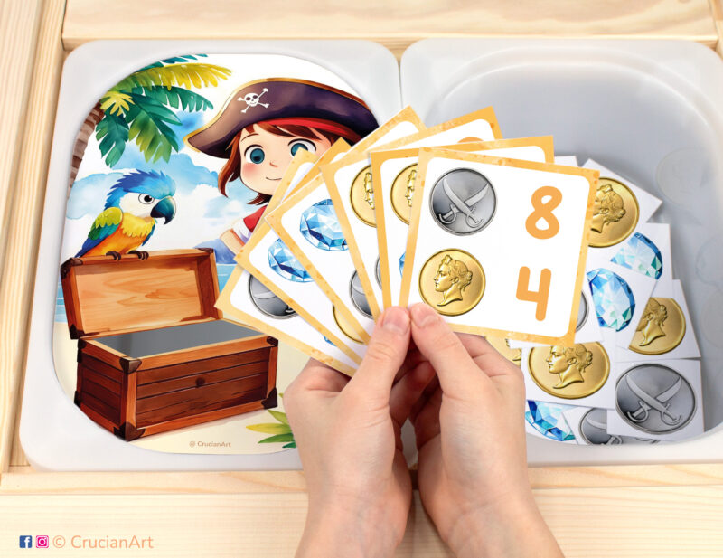 Pirate treasure chest themed pretend play setup for a matching and counting game. Kids' hands holding task cards displaying numerals and gold coins, silver coins, gemstones. Treasure Island Adventure unit printables for toddlers.