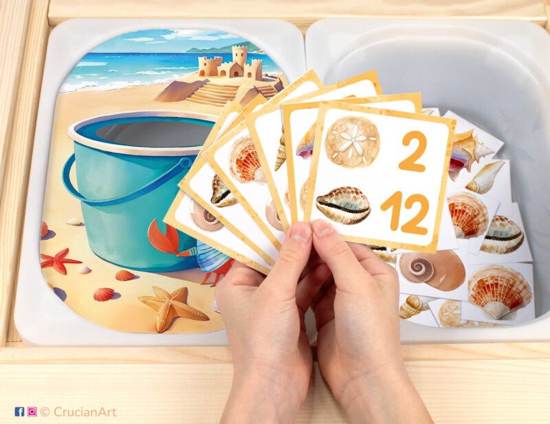 Beach-themed pretend play setup for a matching and counting game. Kids' hands holding task cards displaying numerals and seashells. Summer playtime unit printables for toddlers.