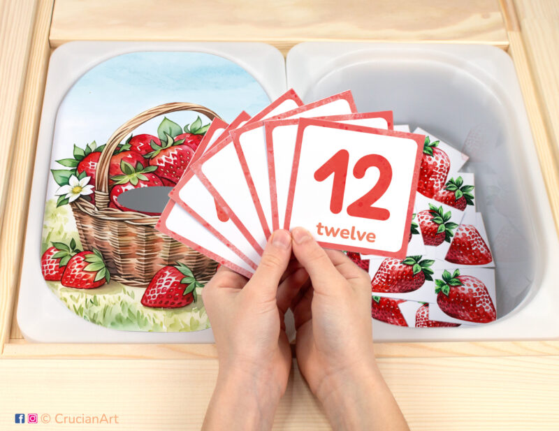 In the garden pretend play setup. Sensory table insert and kids hands holding task cards displaying numerals from 1 to 12. Strawberry unit printable activity for summer season.