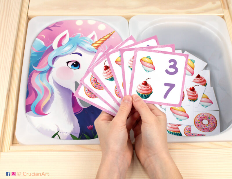 Feed the Magical Unicorn pretend play setup for a matching and counting game. Kids' hands holding task cards displaying numerals and cupcakes, marshmallows, and donuts. Rainbow unicorn printables for toddler girls.