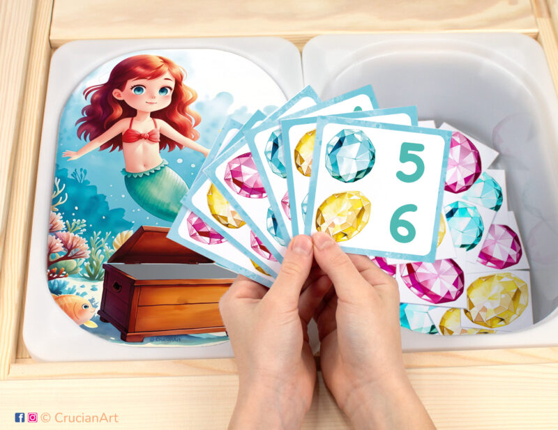 Mermaid treasure chest themed pretend play setup for a matching and counting game. Kids' hands holding task cards displaying numerals and gemstones. Treasure Island Adventure unit printables for toddler girls.