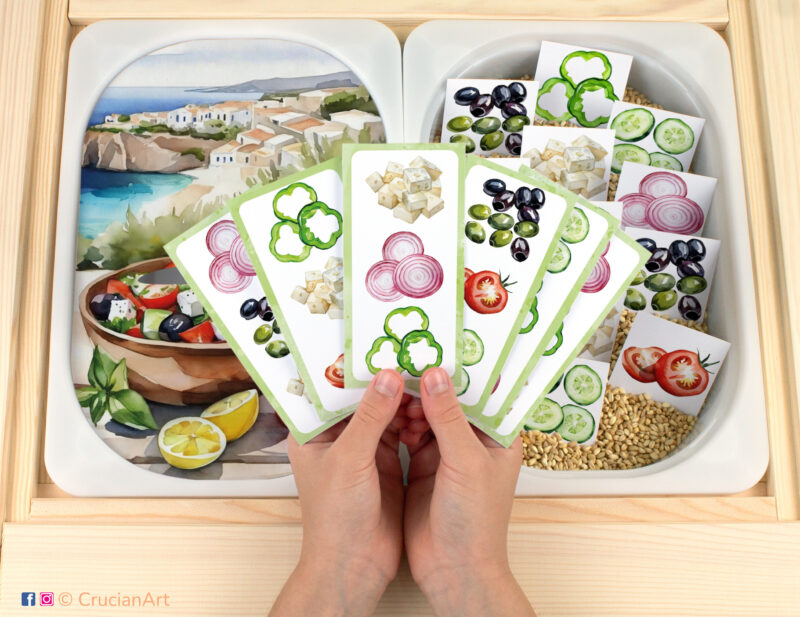 Greek salad making pretend play setup for a matching game. Kids' hands holding task cards displaying tomatoes, cucumbers, green bell pepper, olives, red onion, and feta cheese.. Summer season printables for toddlers.