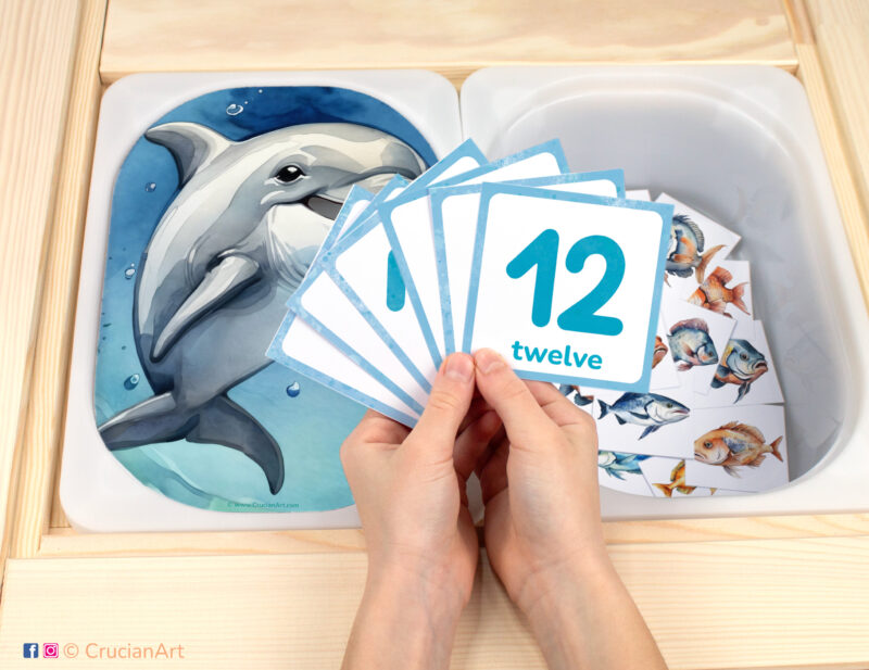 Feed the Dolphin fish pretend play setup. Sensory table insert and kids hands holding task cards displaying numerals from 1 to 12. Printable activity for under the sea animals unit.